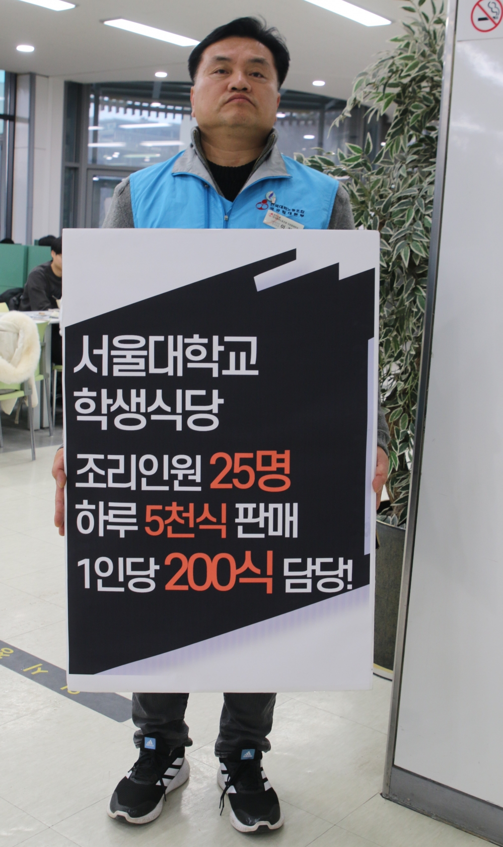 사진설명 시작. 파란 조끼를 입은 남성이 서울대학교 학생식당 조리인원 25명 하루 5천식 판매, 1인당 200인분 감당! 이라고 적힌 피켓을 들고 서 있다. 사진설명 끝.