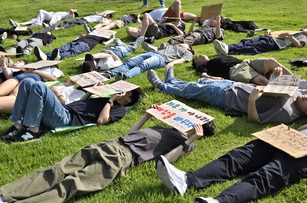 사진 설명 시작. 잔디밭에 학생들이 일제히 누워 있다. 피켓으로 얼굴을 가린 사람도 있고, 피켓 없이 뙤약볕을 정면으로 마주한 사람도 있다. 사진 설명 끝.