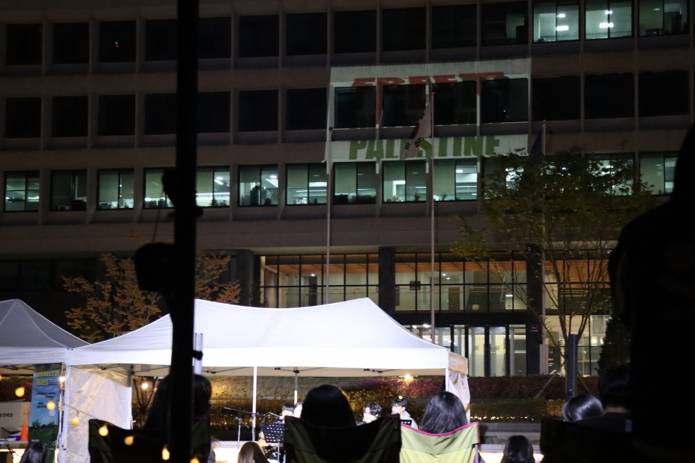 사진설명 시작. 잔디광장에서 축제 공연을 보는 학생들의 뒷모습이다. 학생들 위로 행정관에 빔프로젝터로 띄워진 Free Palestine 문구가 보인다. 사진설명 끝.