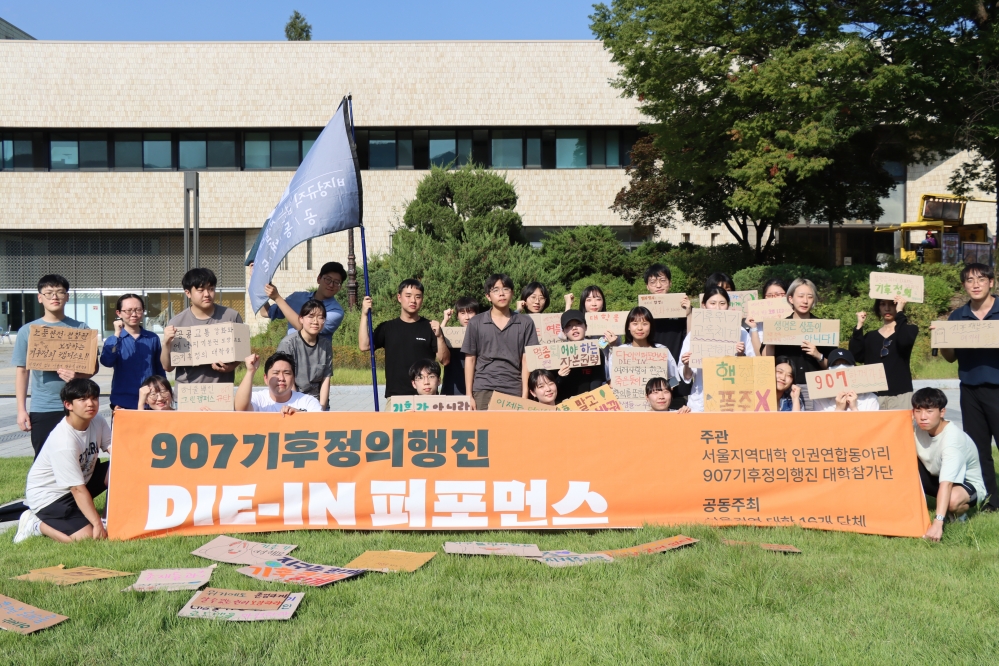 사진 설명 시작. 참가자들이 다이인 퍼포먼스를 끝내고 짧은 기념촬영 시간을 가지고 있다. '907기후정의행진 DIE-IN 퍼포먼스, 주관 서울지역대학 인권연합동아리, 907기후정의행진 대학참가단'이라는 문구가 적힌 현수막을 넓게 펼치고 있다. 사진설명 끝.