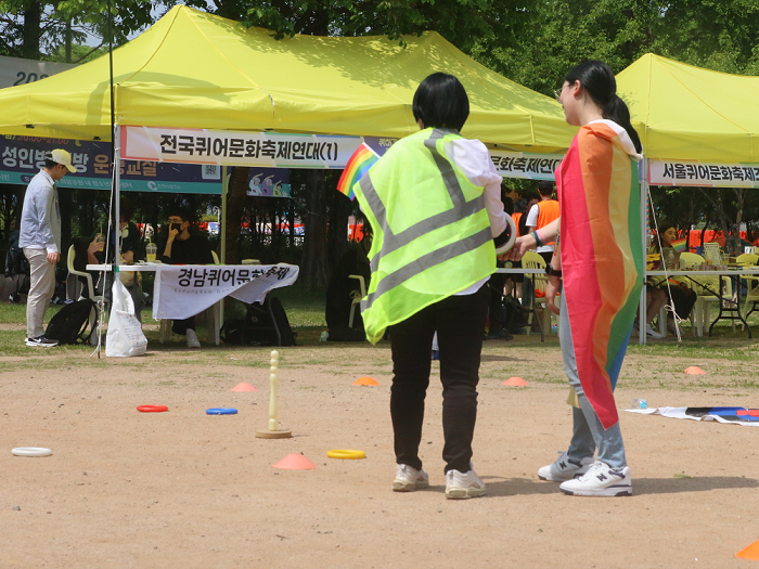 사진 설명 시작. 전국퀴어문화축제연대라 쓰인 노란색 부스들 앞에 고리 던지기 놀이기구가 놓여있다. 색색의 고리와 함께 깃발을 몸에 두른 관계자 두 명이 서있다. 사진 설명 끝.