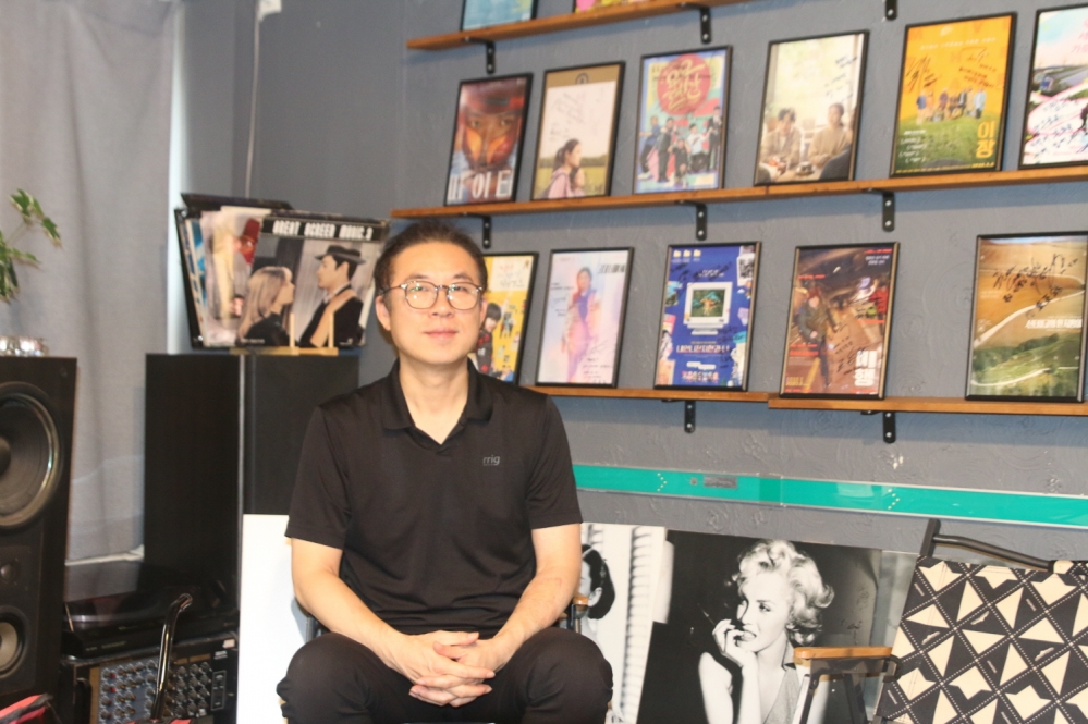 사진 설명 시작. 안경을 쓴 최현준 대표가 영화포스터들이 가득한 벽을 배경으로 두 손을 모은채 정면을 응시하고 있다. 사진 설명 끝.