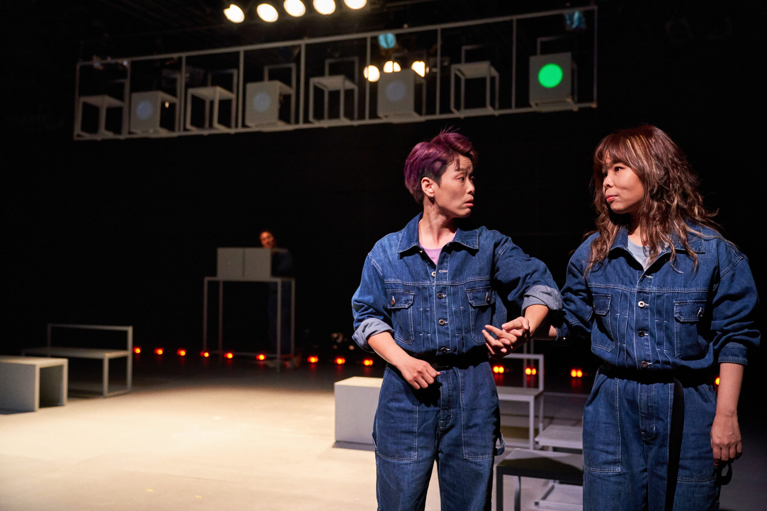 사진 설명 시작. 두 명의 인물이 사진 오른쪽에서 서로 손을 잠고 이야기를 나누고 있다. 두 사람은 모두 청색의 점프수트를 입고 있고, 소매를 팔까지 걷은 차림이다. 왼쪽 사람은 붉은 색 계열의 짧은 머리, 오른쪽 사람은 갈색 긴 머리를 하고 있다. 두 사람 뒤로는 여러 각진 의자가 널부러져 있다. 왼쪽 위에는 가로로 눕힌 사라디 형태의 구조물이 무대 천장에 걸려 있는데, 각 빈칸마다 의자가 한 개씩 놓여 있다. 사진 설명 끝.