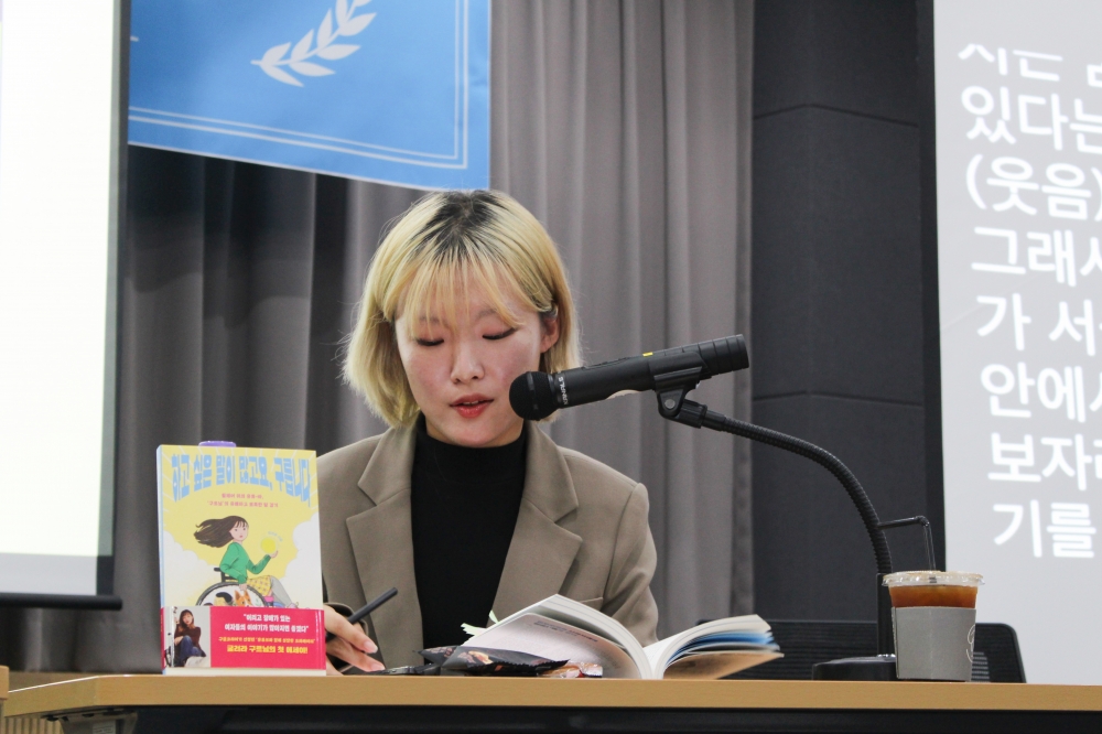 사진 설명 시작. 노란 단발 머리에 검정색 티셔츠, 갈색 자켓을 입고 있는 구르님이 마이크에 대고 발언하는 모습이다. 구르님 앞에는