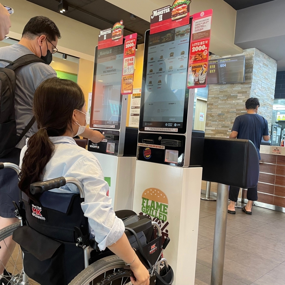 사진 설명 시작. 햄버거 패스트푸드 가게 키오스크 앞에 휠체어를 탄 머리카락의 길이가 긴 사람이 앉아있다. 이 사람은 키오스크를 위로 올려다보고 있다. 옆에서는 휠체어를 타지 않은 사람이 서서 키오스크를 이용하고 있다. 사진 설명 끝.