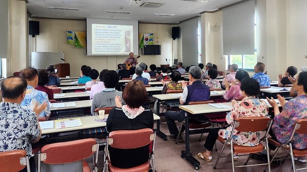 사진 설명 시작. 강의실의 모습이다. 맨 앞 쪽에는 PPT 스크린이 띄워져 있다. 기타를 든 노인과 박수 치며 호응하는 노인들의 모습이다. 이들은 웰다잉 프로그램에 참여하고 있다. 대략 30명 정도의 인원이 강의를 수강하고 있는 모습이다. 사진 설명 끝.