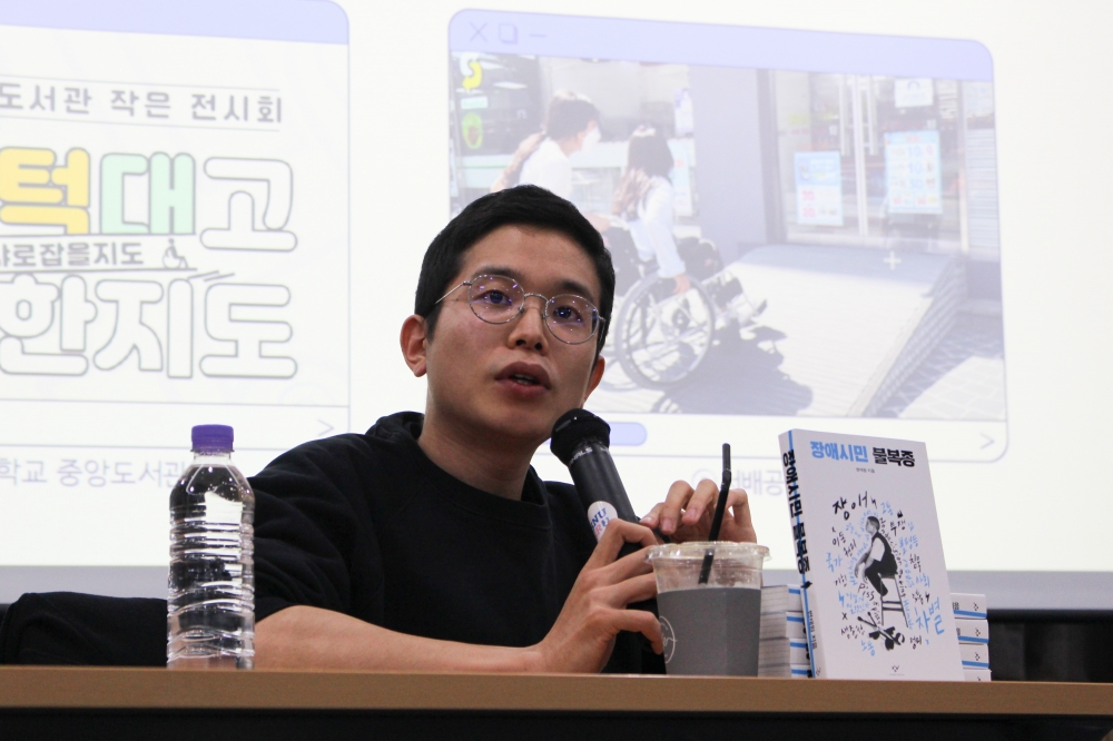 사진 설명 시작. 변재원 작가는 짧은 머리에 회색 동그란 안경을 쓰고 검정색 티셔츠를 팔꿈치 위로 걷어 올린 채로 마이크를 들고 앉아 있다. 그의 앞에는 왼쪽부터 물이 든 페트병, 일회용 플라스틱 커피 잔, 장애시민 불복종 책이 놓여 있다. 변재원 작가의 뒤편으로는 ppt화면이 나오고 있다. 사진 설명 끝.