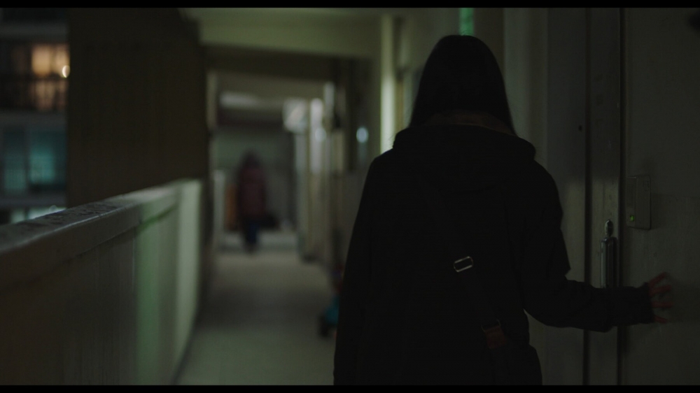 사진 설명 시작. 복도식 아파트에서 수경과 이정이 집으로 가고 있다. 가만히 서있는 이정의 뒷모습이 보인다. 이정은 복도 오른편에 붙어 오른 손으로 벽을 짚고 앞을 바라보고 있다. 흐린 초점 사이로 앞서가는 수경의 앞모습이 보인다. 사진 설명 끝.