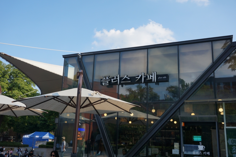 사진 설명 시작. 플러스카페의 외부 사진이다. 푸른 하늘 앞으로 투명한 유리창이 외벽에 붙은 건물이 있고,