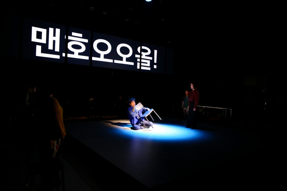 사진 설명 시작. 어두운 무대 위에 남자 배우가 의자를 끌어앉고 주저앉아 있다. 세 화면으로 분할된 배경의 스크린에는, 맨호오오올! 이라고 적힌 거대한 자막이 볼드체로 띄워져 있다. 사진 설명 끝.