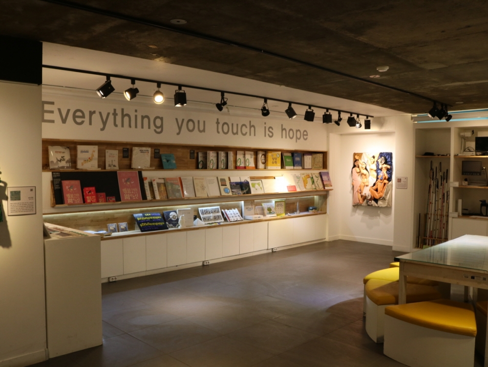사진 설명 시작. 흰색 전시장 벽에 점자책과 교재들이 세 열로 진열돼 있다. 진열장 위쪽에는 다음과 같은 문구가 영어로 크게 적혀 있다. 당신이 만지는 모든 것은 희망입니다. 사진 설명 끝.
