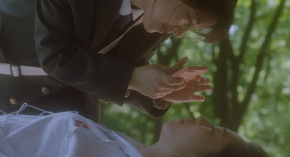 사진 설명 시작. 녹색 숲을 배경으로, 세미가 누워 있는 하은을 휴대폰 카메라로 찍고 있다. 하은은 눈을 감고 있다. 사진 설명 끝.