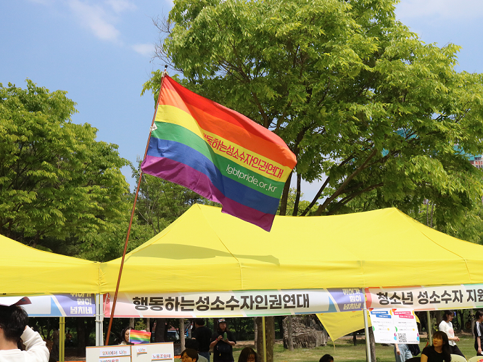 사진 설명 시작. 울창한 초록빛 나무들을 배경으로 행동하는성소수자인권연대 부스가 설치돼 있다. 노란 천막으로 덮인 부스 위에는 행동하는성소수자인권연대라고 쓰인 무지갯빛 깃발이 휘날리고 있다. 사진 설명 끝.