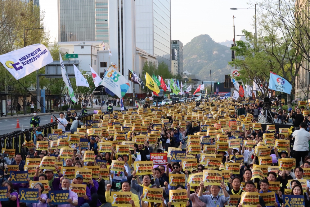 사진 설명 시작. 시민들이 한데 모여 피켓을 들고 구호를 외치고 있다. 여러 단체의 깃발이 흔들리고 있고, 피켓에는