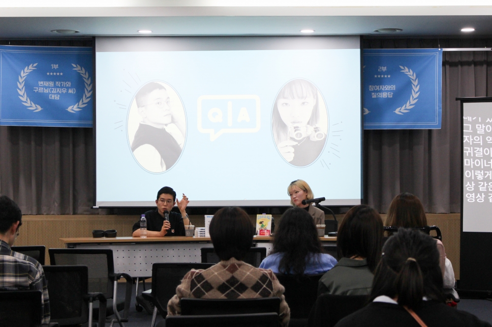 사진 설명 시작. 사진 속에는 변재원 작가와 구르님이 맨 앞에 나란히 앉아 있다. 강연을 듣는 사람들의 뒷모습이 보인다. 변재원 작가는 대담에서 발언을 하면서 한쪽 손을 펼치고 얼굴 옆에 올리고 있다. 구르님과 변재원 작가의 뒤편으로는 프로젝터에 ppt가 나오고 있다. ppt에는 왼쪽에는 변재원 작가의 사진이, 오른쪽에는 구르님의 사진이 있고 그 사이에 Q&A라고 적혀 있다. 빔 프로젝터 뒤로 행사 현수막이 나와있고 왼쪽에는 1부 변재원 작가와 구르님(김지우 씨) 대담이라고 적혀 있고 오른쪽에는 2부 참여자와의 질의응답이라고 적혀 있다.