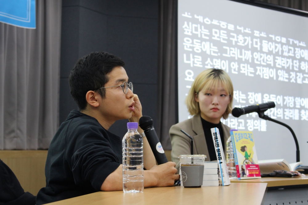 사진 설명 시작. 변재원 작가가 마이크를 들고 이야기하는 옆모습이 나와 있다. 그의 앞에는 물이 든 페트병이 놓여 있다. 변재원 작가의 오른쪽 뒤편으로 구르님이 변재원 작가의 이야기를 경청하는 모습이 나와 있다. 구르님의 뒤편으로는 문자 통역 화면이 나오고 있다. 변재원 작가와 구르님 사이에 두 분이 각자 쓰신 책들이 세워져 있다. 사진 설명 끝.