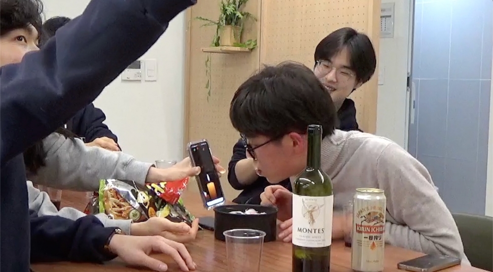 사진 설명 시작. 와인과 맥주, 과자를 책상 한가운데 두고 서너명의 남자들이 웃고 있다. 가장 앞에 있는 남자가 생일인듯 휴대폰 화면으로 보이는 초를 끄는 시늉을 하고 있다. 사진 설명 끝.