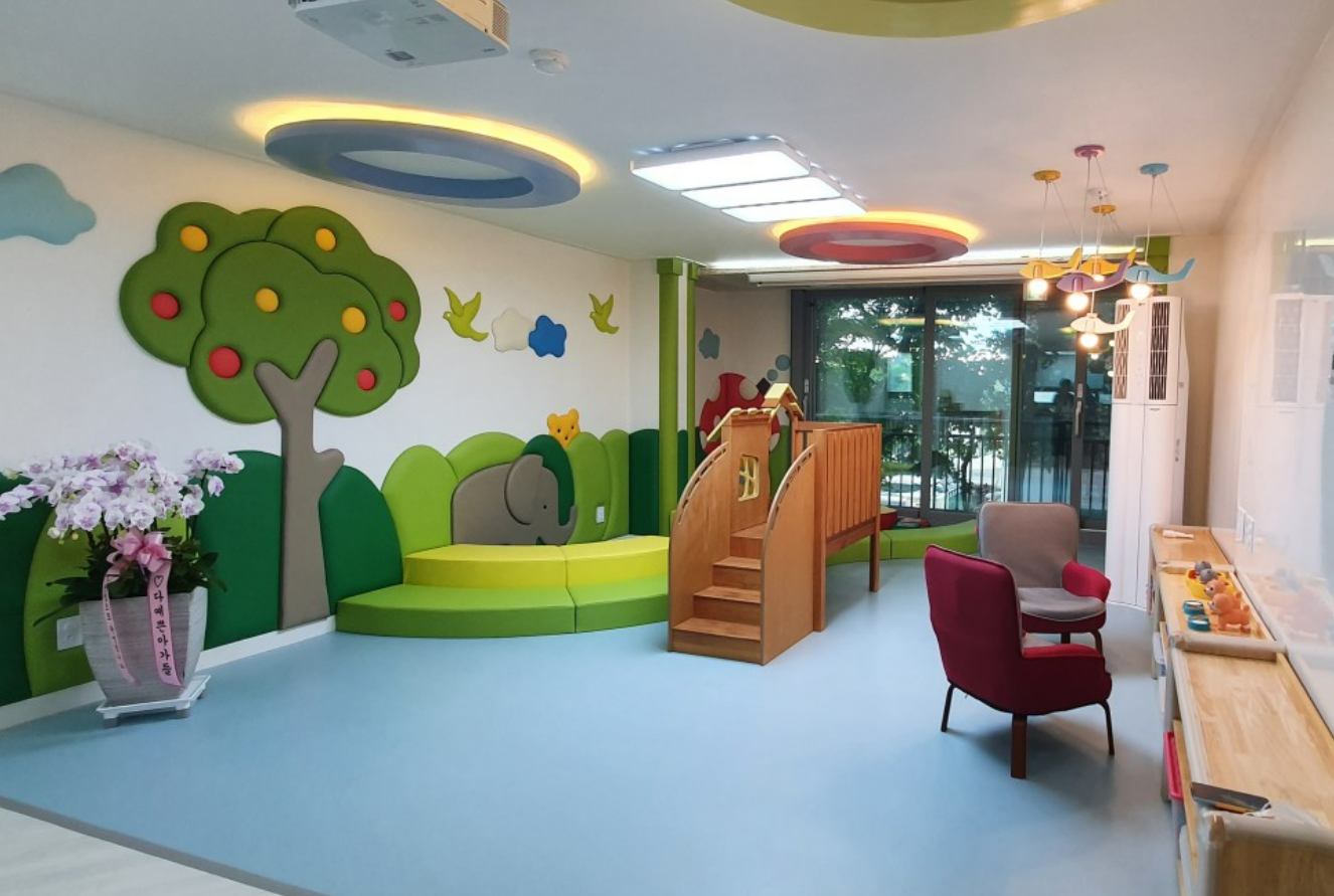 사진 설명 시작. 아파트의 거실이다. 바닥에는 아이들을 위해 푹신한 매트가 깔려있다.한쪽에는 아이들이 놀 수 있는 장난감들이 놓여있고 거실 중앙에 미끄럼틀이 놓여있다. 벽면에는 코끼리, 나무, 새와 구름이 그려져있다. 사진설명 끝.