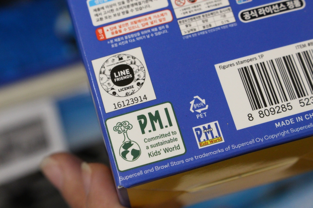 사진 설명 시작. 장난감 포장재 뒷면의 한쪽 귀퉁이가 확대돼 있다. 흰색 배경에 진한 초록색 지구 아이콘이 그려져 있다. 옆에는 PMI라는 회사명이 크게 적혀 있다. 사진 설명 끝.