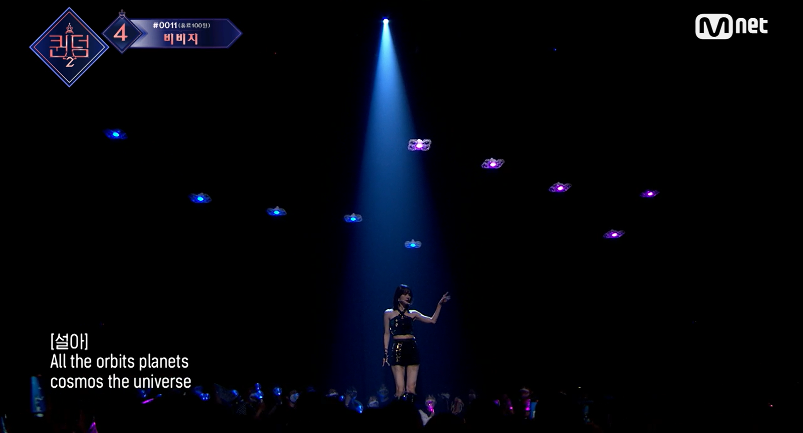 사진 설명 시작. 사진의 왼쪽 위에 방송 프로그램의 로고가 있다. 우주소녀 멤버인 설아가 무대 중앙에서 공연하고 있는 모습이다. 머리 위로 드론들이 행성처럼 떠 다닌다. 무대 중앙을 기준으로 왼쪽에는 파란색을 띠는 드론이, 오른쪽에는 핑크색을 띠는 드론이 날아다닌다. 무대 아래에는 팬들이 응원봉을 가지고 무대를 지켜보고 있다. 사진의 왼쪽 아래에 노래의 가사가 적혀 있다. 사진 설명 끝.