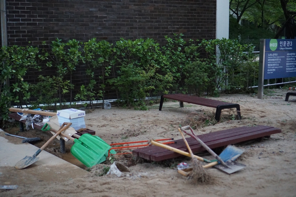 사진 설명 시작. 세 개의 벤치가 흙에 묻혀 있고 벤치 위에는 삽과 빗자루가 있다. 벤치 뒤에는 수목과 인문관 3이라고 적힌 팻말이 있다. 사진 설명 끝.