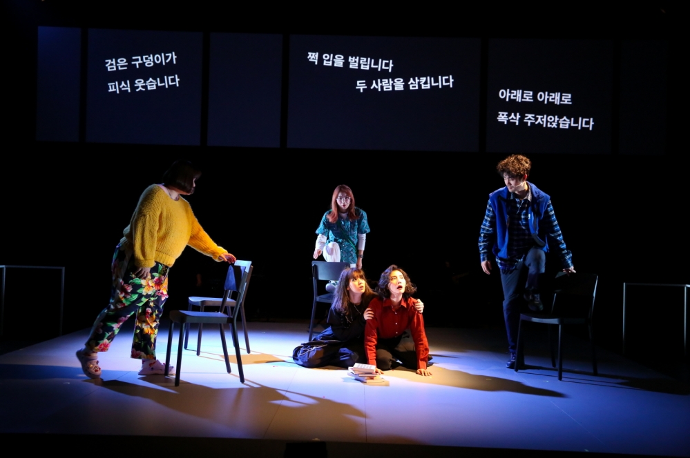 사진 설명 시작. 어두운 무대 위에 다섯 명의 배우가 있다. 두 명의 배우는 절망한 듯 바닥에 엎어져 있고, 세 명의 배우는 각자 의자를 들고 두 배우를 둘러싸고 위협하듯 서 있다. 세 화면으로 분할된 배경의 스크린에는 각각 검은 구덩이가 피식 웃습니다, 쩍 입을 벌리고 두 사람을 삼킵니다, 아래로 아래로 폭삭 주저앉습니다, 라는 자막이 쓰여 있다. 자막은 주인공들의 절망적 심리를 반영하듯 제멋대로 기울어져 있다. 사진 설명 끝.