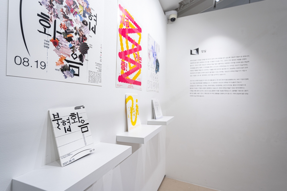 사진 설명 시작. 암실의 제3회 기획전 『춤추는 사람들은 안개 속에서』에 전시된 암실의 지난 전시 도록들이다. 사진의 가장 왼쪽에서부터 2021년 8월에 진행한 『불협화음』 전시의 포스터가 붙어 있고 그 아래 하얀 바탕에 검은 글씨로
