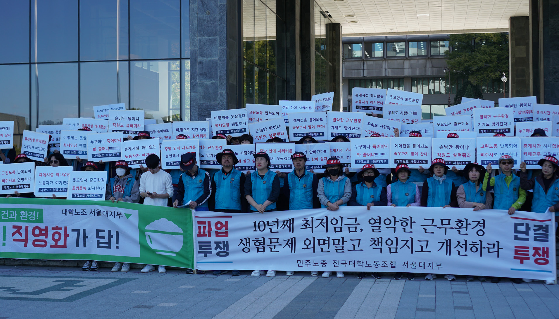 사진 설명 시작. 행정관 앞 파란색 조끼를 입은 노동자들이 열을 이뤄 서있다. 맨 앞 줄 사람들이 들고 있는 플랑에는