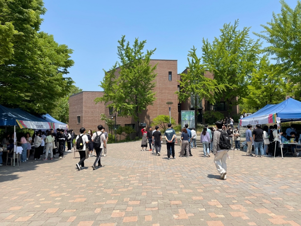 사진 설명 시작. 가운데에 미술대학 건물이 보인다. 중앙 광장을 기준으로 좌우에 파란색 부스들이 설치되어 있다. 각 부스 안에서는 학생들이 음식을 만들거나 게임을 진행하고 있고 그 앞에 참여하려는 학생들이 줄을 서 있다. 가운데에서 구경하는 학생들도 여럿 있다. 사진 설명 끝.