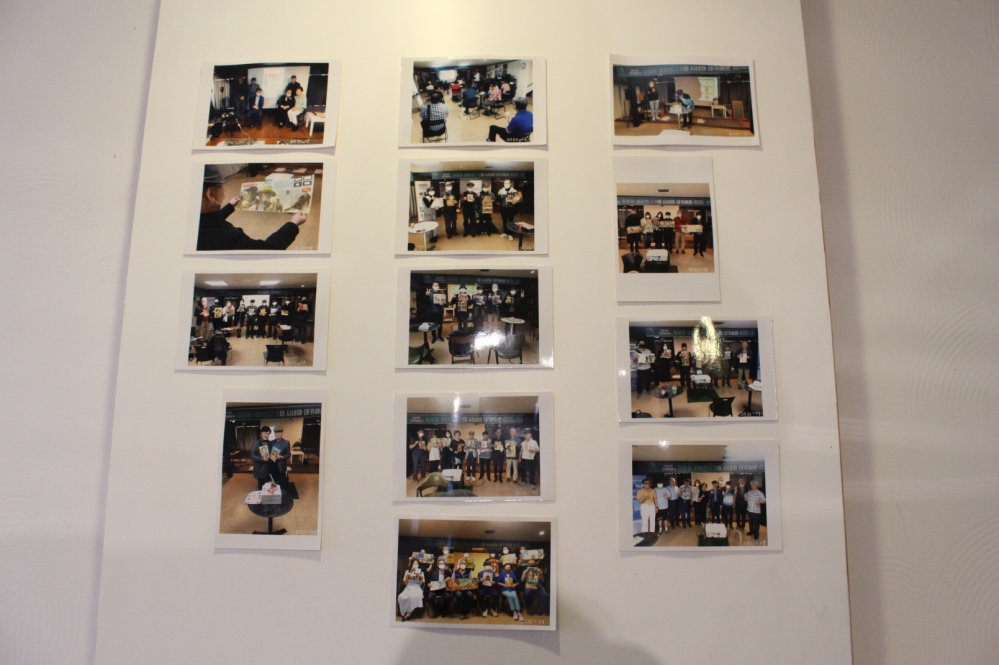 사진 설명 시작. 흰 벽 위에 사진들이 여러장 붙어있다. 각각의 사진은 단체사진이다. 카메라를 바라보고 포즈를 취하는 사람들의 모습이 보인다. 사진 설명 끝.