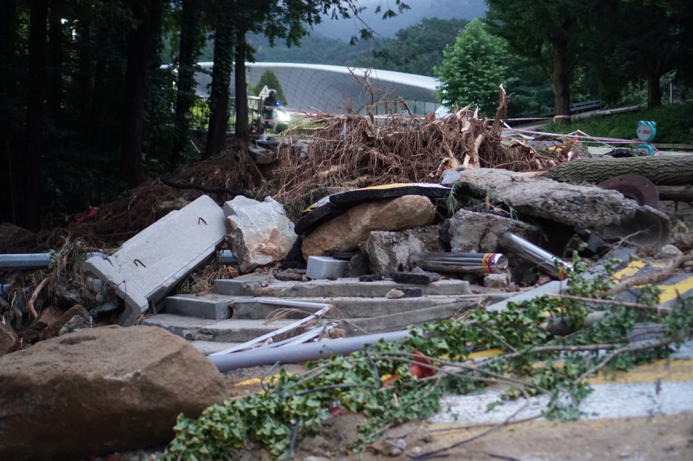 사진 설명 시작. 부러진 나무와 나뭇가지, 망가진 아스팔트 잔해, 콘크리트 잔해 등이 바닥에 뒹굴고 있다. 뒤에는 버들골 풍산마당이 있다. 사진 설명 끝.