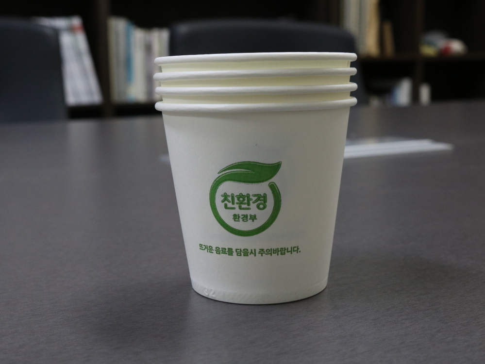 사진 설명 시작. 네 개의 종이컵이 회색 테이블 위에 겹쳐진 상태로 놓여 있다. 종이컵은 흰색이고 종이에 적힌 글씨는 초록색이다. 종이컵 가운데에