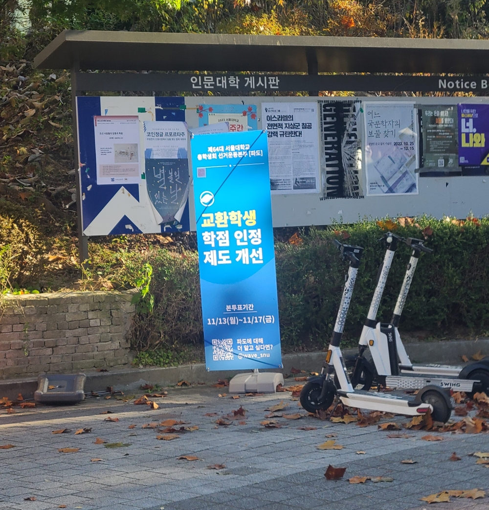 사진 설명 시작. 인문대학 게시판 앞에 제64대 총학생회 선거운동본부 ‘파도’의 공약 홍보용 판넬이 설치돼있다. 게시된 판넬 중앙에는 교환학생 학점 인정 제도 개선이 써있다. 판넬 하단에는 본투표기간 및 파도의 큐알코드가 명시돼있다. 판넬 옆에는 킥보드가 3대 세워져있다. 사진 설명 끝.