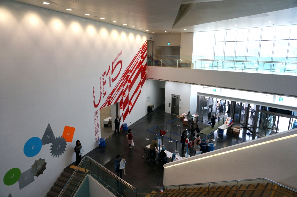 사진 설명 시작. 지난 11월 열린 아트북‧독립출판 페어 ‘언리미티드 에디션’ 현장의 사진이다. 계단 위에서 아래로 행사 현장을 찍은 사진으로, 왼쪽 흰 벽에는 행사 제목이 빨간 글씨와 함께 디자인되어 써있다. 사진 오른쪽으로는 행사장으로 입장하는 여러 개의 문이 있어 많은 사람들이 들어오고 있다. 사진 설명 끝.