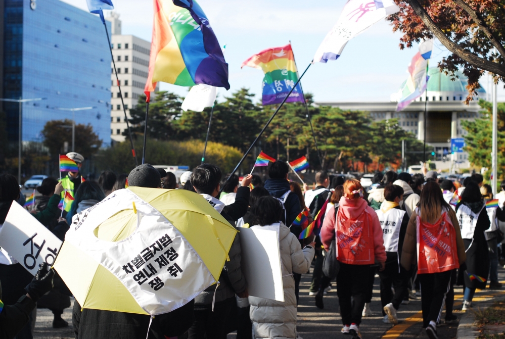 사진 설명 시작. 무지개 색 깃발을 든 사람들의 행진이 사진 오른쪽의 국회의사당을 향하고 있다. 왼쪽 하단에는 차별 금지법 연내 쟁취라는 문구가 쓰인 우산을 쓰고 걸어가는 시민의 뒷모습이 있다. 사진 설명 끝.