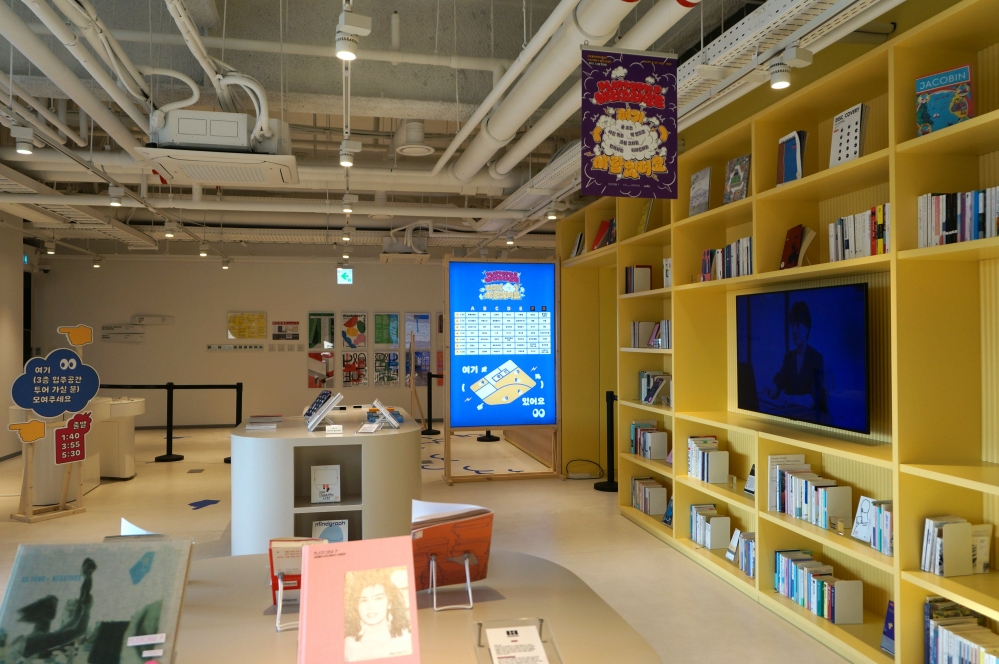 사진 설명 시작. 플랫폼P에서 진행된 행사 ‘여기 ( ) 사람 있어요’ 현장이다. 오른쪽에는 노란색 책장이 벽의 역할을 하고 있다. 가장 중앙에는 큰 TV가 있고, 그 주위로 책장에 여러 책이 꽂혀 있다. 중앙에는 책상 위에 여러 책이 전시돼있고, 그 뒤로는 스크린에