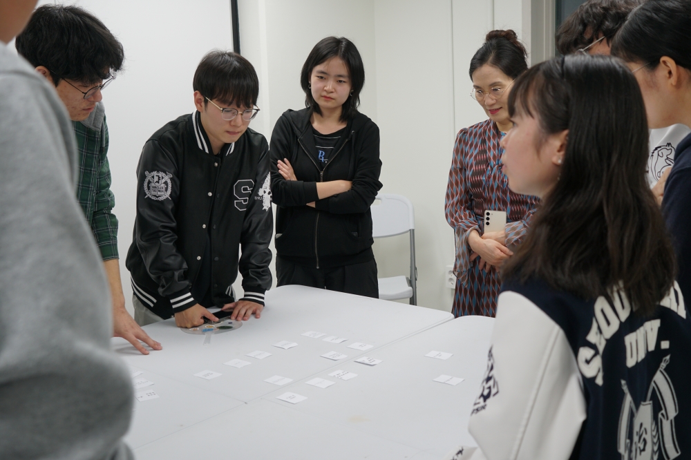 사진 설명 시작. 글귀가 적힌 카드를 늘어놓은 흰 테이블 주위에 학생들이 서있다. 학생들의 시선은 테이블 위 카드를 향해있다. 사진 설명 끝.