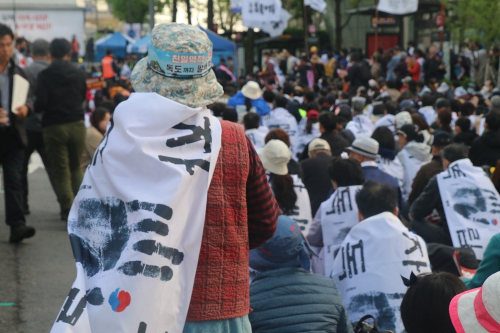 사진 설명 시작. 거리에 시위자들이 줄지어 앉아 있다. 사진의 초점은 가까이에 서 있는 한 시위자에 맞춰져 있다. 초점이 맞은 시위자는 카메라에 등을 돌리고 있다. 시위자들이 망토처럼 두른 깃발에는 안중근 의사의 손도장과
