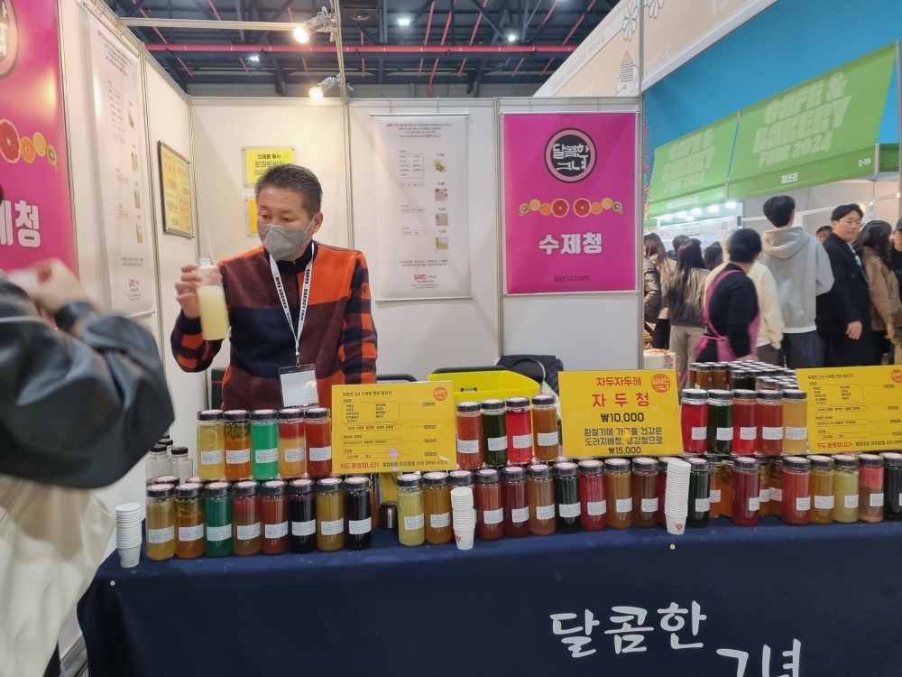 사진 설명 시작. 카페베이커리 부스 중 과일 수제청을 팔고 있는 부스다. 부스담당자가 수제청을 탄 음료를 들고 있다. 사진 설명 끝.