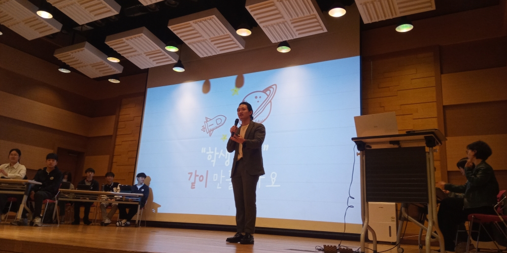 사진 설명 시작. 파란색 스크린이 띄워진 무대에 한 남성이 마이크를 쥐고 서있다. 무대에 서있는 사람 기준 오른쪽에 학생들이 책상 뒤에 앉아 있고 왼쪽에는 두 조교가 화면을 바라보고 있다. 사진 설명 끝.