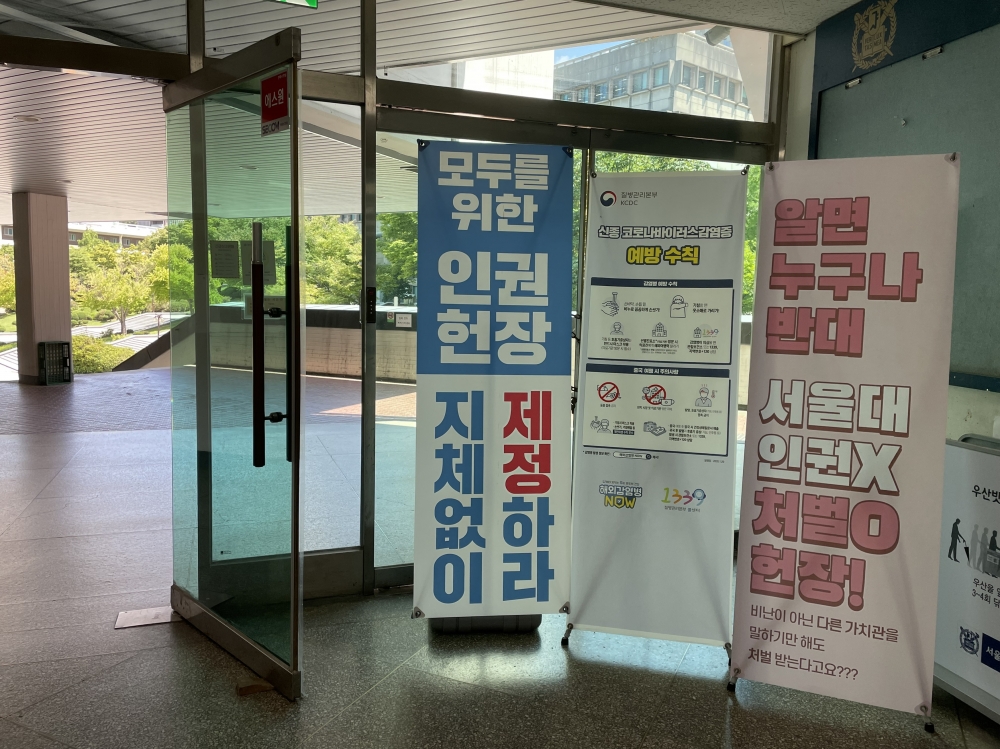 사진 설명 시작. 학생회관 2층 건물에 배너 3개가 놓여있다. 가장 왼쪽에는 ‘모두를 위한 인권헌장 지체없이 제정하라’라고 적혀있으며, 제정이라는 글자가 빨간색으로 칠해져있다. 이 배너는 파란색과 흰색이 주로 사용됐다. 가운데 배너의 경우 신종코로나 바이러스 감염증 예방 수칙이 적혀있다. 작은 그림과 글씨로 수칙을 설명하고 있다. 가장 오른쪽에는 ‘알면 누구나 반대 서울대 인권 X 처벌 O 헌장! 비난이 아닌 다른 가치관을 말하기만 해도 처럽다는다고요???’라고 적혀있다. 이 배너는 분홍색 톤이다. 사진설명 끝.