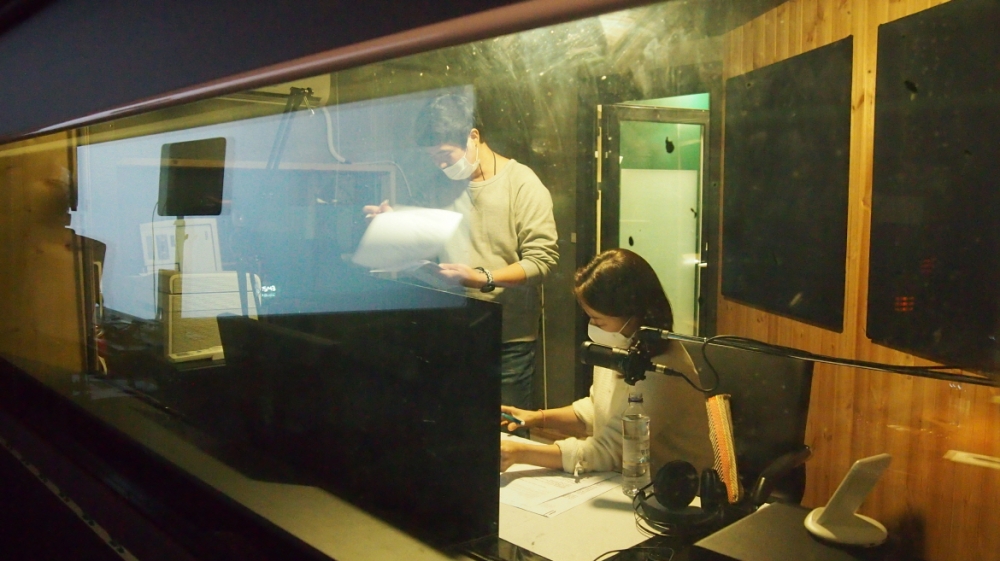 사진 설명 시작. 배리어프리 영화 녹음현장이다. 녹음실 왼쪽에서는 대본을 손에 든 남자 성우가 대사를 녹음한다. 오른쪽에는 앉아 있는 여자가 성우가 대사를 녹음한다. 사진 설명 끝.