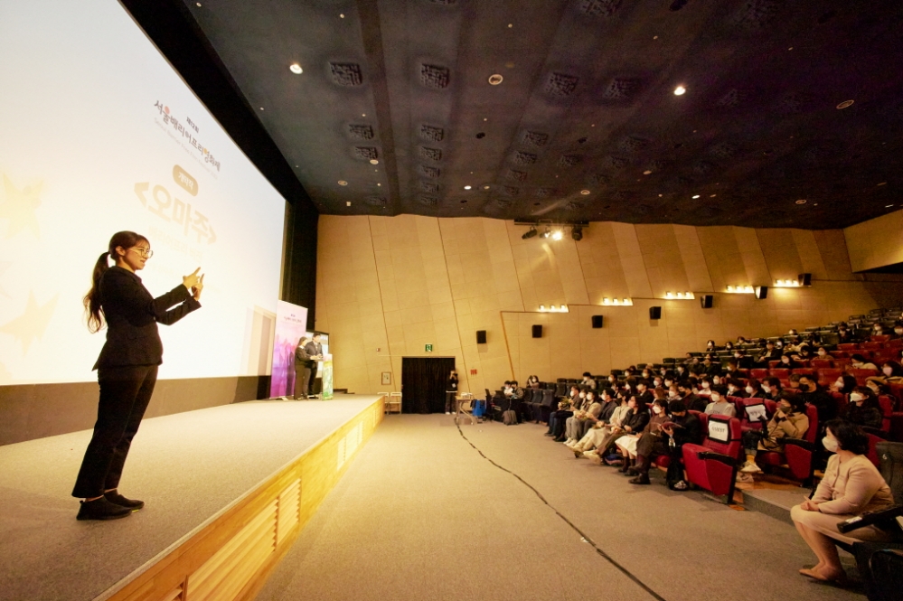 사진 설명 시작. 서울배리어프리영화제 개막식이다. 커다란 극장의 스크린에서 상영작이 소개되고 있다. 스크린 앞 무대에서 수어통역사가 소개 내용을 통역하고 있다. 관객들이 극장에 가득 앉아 있다. 사진 설명 끝.