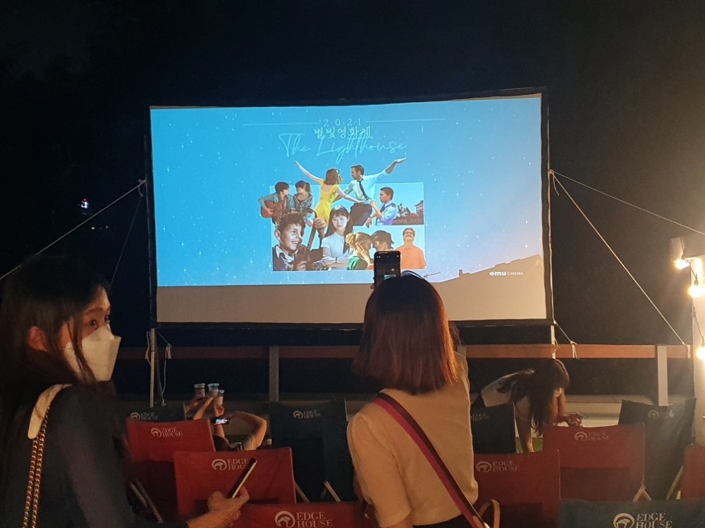 사진 설명 시작. 에무시네마에서 열린 《라라랜드》 야외상영회 장면이다. 야외 루프탑에 스크린이 설치돼 있고 스크린 앞에서 관객들이 기념사진을 촬영하고 있다. 스크린에는