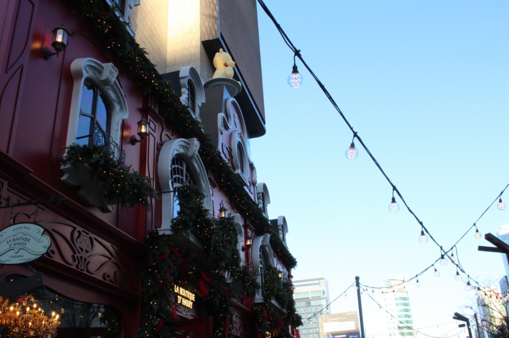 사진 설명 시작. 파란 하늘을 배경으로 빨간 백회점 크리스마스 팝업스토어가 서있다. 팝업스토어 앞 전깃줄에는 여러개의 전구가 달려있다. 사진 설명 끝.