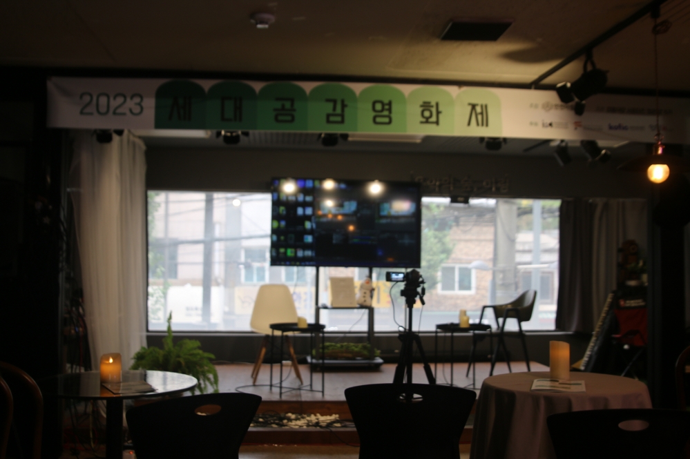 사진 설명 시작. 「2023 세대공감영화제」 녹색과 흰색이 섞인 현수막이 걸려있는 무대 아래 두 개의 흰색 의자가 있고 그 앞에 녹화용 카메라가 있다. 무대 아래에는 원형의 테이블과 의자로 이뤄진 관객석이 있다. 사진 설명 끝.