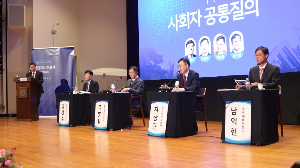 사진 설명 시작. 가장 왼쪽에 사회자가 교단 뒤에 서 있고, 그 옆으로 이철수, 유홍림, 차상균, 남익현 후보자가 차례대로 무대 위 마련된 책상과 의자에 앉아 있다. 책상 앞에는 각 후보의 이름과