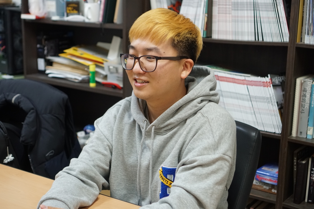 사진 설명 시작. 서울대저널 편집실에 김철주 선관위장이 앉아있다. 노란 머리에 안경을 쓴 그는 회색 후드티를 입고 있다. 얼굴에는 살짝 미소를 머금고 있다. 의자에 앉아있고 손을 바로 앞 테이블 위에 놓고 있다. 사진 설명 끝.