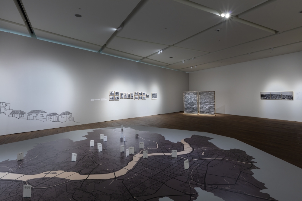 사진 설명 시작. 『뮈에인, 내 마음속의 오목렌즈』의 전시 전경이다. 바닥에는 서울의 지도가 그려져있고 그 위에 각 구의 이름이 써있는 하얀색 팻말이 서있다. 그 뒤로는 흰색 전시장 배경에 단층집의 그림의 그려져있고, 그 오른쪽으로는 흑백 사진 작품들이 줄을 지어 전시되어 있다. 사진 설명 끝.
