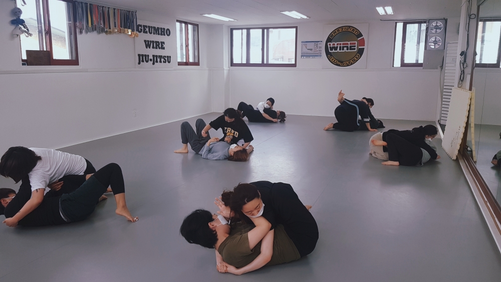 사진 설명 시작. 실내 체육관에서 여성들이 주짓수를 배우고 있다. 각각 두 명씩 짝을 이뤄 총 여섯 쌍이 서로 몸을 격하게 맞대고 동작을 연습하고 있다. 모두 한 명은 누워 있고 다른 한 사람이 그 위에 올라가 있는 자세로 서로 공격하고 방어하는 모습을 하고 있다. 가장 앞쪽에 위치한 두 사람 중 한 명은 밑에 있는 사람의 위에 올라 상대방의 상체를 두 팔로 힘껏 감아 안고 있고, 밑에 있는 사람은 제압을 풀기 위해 두 손을 모은 자세로 상대방의 머리와 목 부분을 밀어내고 있는 것으로 보인다. 사진 설명 끝.